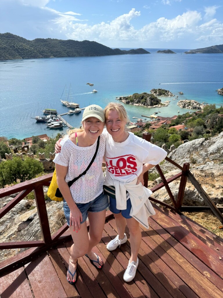 Two women stand arm in arm smiling with the view of the sea behind them